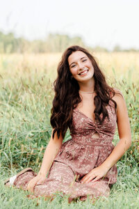 A high school senior sits in the grass and smiles for the camera. She is wearing a long floral dress.