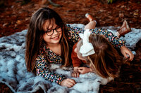 Two little girls lay on a blanket on their stomachs looking at each other and giggling in this family fall photography session/