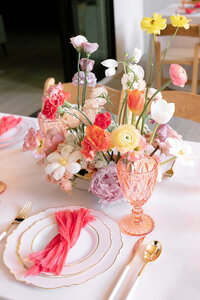 Brightly colored  table place setting with florals and orange napkin and pink glass