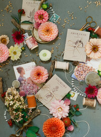 A 35mm film photograph showcasing a styled flat lay from a florist branding session. The image features an artful arrangement of vibrant flowers, including dahlias, hydrangeas, and berries, paired with vintage seed packets, spools of ribbon, delicate pins, scissors, and a small brass tray. The soft, natural tones and intricate details highlight the craftsmanship and creativity of floral design, creating a visually stunning composition perfect for storytelling and brand elevation.