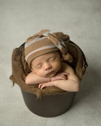 Newborn boy with tan and white sleepy hat sleeping in a pail.