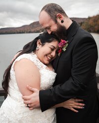 A smiling bride leans into her groom's chest as they snuggle by the water