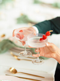 Two coupe glasses cheersing with raspberries in drinks