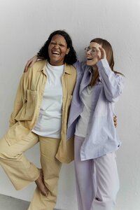Two feminine presenting people stand against a white wall, one wearing a yellow monochromatic outfit, and the other a similar purple outfit. They have their arms wrapped around each other, and they are both smiling happily.