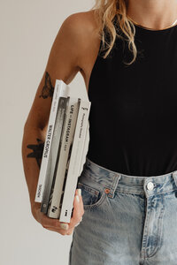 A woman in light denim jeans and a tucked-in black vest top. The image captures her torso and hips, with one arm holding a stack of books. Some of her blonde hair cascades over her shoulder.