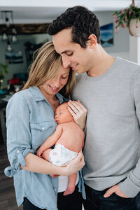 A couple stands closely together, holding their newborn baby. The mother gently cradles the baby, while both parents look down lovingly at their child.