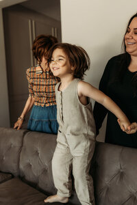 A young child gleefully jumps on the couch while holding hands with their mom and grandmother, all smiling during a playful in-home family session in Scottsdale, Arizona.