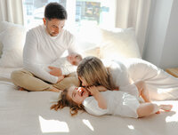 mom tickling toddler on bed while dad looks on holding newborn baby