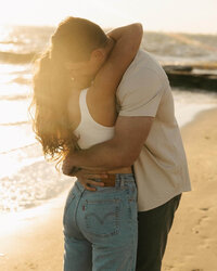 A happy engaged couple embraces on a beach at sunset in jeans and white tops