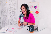 Jessi sitting in front of a microphone talking in her podcast. She is sitting a desk.