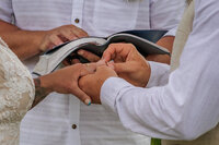 Bride and groom with the groom putting the ring on the brides hand with the officiant holding a bible right behind them.
