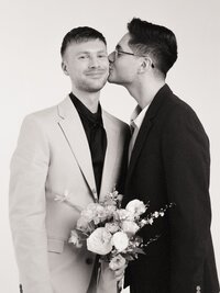 Black and white film portrait of two grooms in a studio setting, one holding a bouquet and smiling as his partner kisses him on the cheek — captured by a film wedding photographer