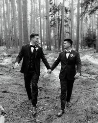 Photo of two grooms walking through a forest while holding hands and smiling at each other taken by Maine wedding photography