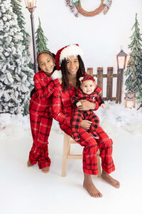 A family of threec children dressed in matching red plaid pajamas, poses in a festive winter backdrop at a Jacksonville portrait studio. The eldest brother is seated in the center holding a baby, while another child stands beside them. Snow, lanterns, and evergreen trees enhance the cozy holiday setting.