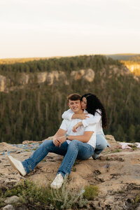 A couple sitting at a cliff's edge. The man is smiling at the camera while the woman kisses him on the cheek.