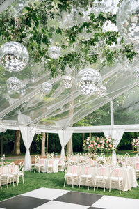 A spacious outdoor tent at Boxwood Estate, set up for a wedding with elegant decor. The tent is large enough to accommodate up to 350 guests, with long banquet tables arranged neatly underneath sparkling string lights. The open sides offer views of the surrounding lush greenery, and floral arrangements and candles add a romantic touch to the ambiance. The setting is both sophisticated and welcoming, perfect for a grand celebration.