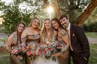 A group of five people, four women in gold dresses and a man in a dark suit, smile while holding flower bouquets. Captured by a Colorado Photographer, they are outdoors with trees in the background and sunlight filtering through.