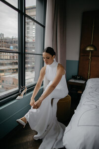 Bride sitting by window in downtown hotel room putting heel on