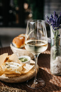 A glass of white wine is on a wicker table. Beside it, there's a wooden board with snacks wrapped in paper and a small bowl of dip. A basket with bread and a vase with purple flowers are in the background.