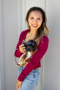 Portrait of a smiling woman photographer holding a Canon camera with a floral strap, captured by Ellobelle Photography Family Photographer