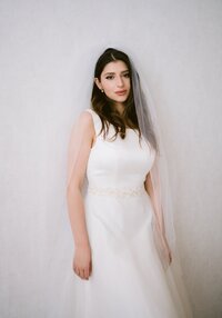 A bride with a white dress and a veil standing by a white wall.