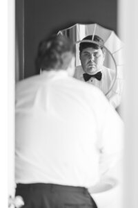 Black and white image of groom adjusting his tie, captured through his reflection, showcasing quiet anticipation before his wedding day begins.