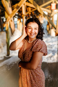 Senior photos of a girl at Central Florida's famed Cocoa Beach Pier. She is posing on a sandy wooden pier while seated wearing a pink and white floral patterned dress. 