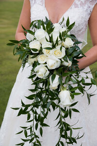 Bride holds a cascading bouquet of white roses and lush greenery, complementing her lace wedding gown at a Jackson Hole celebration.
