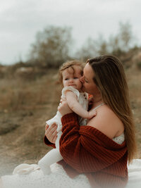 A mama cuddling and kissing her baby girl during a natural light motherhood session in a rustic Southern California field.