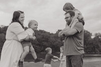 A black and white photo of a mother holding her youngest son and a father carrying another son on his shoulders while their other two children play in the background at Winged Deer Park in Johnson City, TN