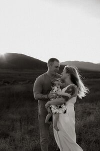 Family of three embracing outdoors, with parents holding their child close in a candid black and white moment.