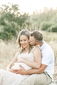 Pregnant woman in light neutral floral dress sitting down with man behind her. They are both holding her baby bump and the man is kissing her on the cheek. Portland Maternity Outdoor Photography Session