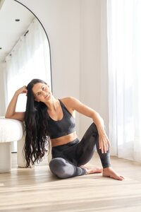 Sarah, a professional yoga mentor, relaxes in a casual seated pose, exuding calmness and approachability in a serene studio environment with natural light filtering through sheer curtains.