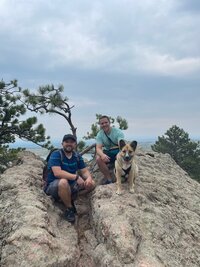 two men sitting on rocks with dog and pine trees in background