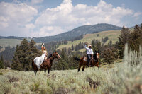 Bride and groom ride horses through an open sagebrush field in Montana during their adventure elopement. The bride wears a flowing white wedding dress, raising her hand in joy, while the groom, dressed in a cowboy hat and western attire, smiles as they explore the rolling hills and mountain backdrop.