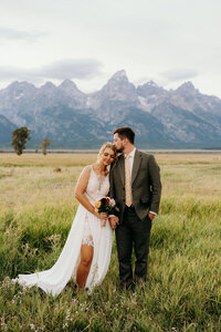 bride and groom standing outside in a field while bride leans her head on groom's shoulder with mountains in the background