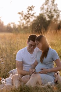 Paar sitzt in der Sonne auf einer Wiese, beide halten den Babybauch der Frau