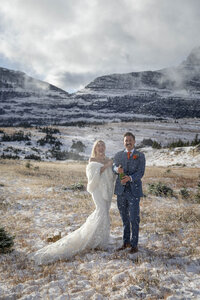 Bride and groom celebrate their winter elopement in Glacier National Park, Montana, popping champagne in a snow-dusted alpine meadow. The bride wears an off-the-shoulder lace wedding dress with a cozy shawl, while the groom, dressed in a navy suit, smiles as snow flurries swirl around them.