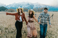 Grand Tetons family photographer Adriana Montoya Photo. A 2 girls and 1 guy, presumably family, holding hands and walking towards the camera. Behind them are the Grand Tetons.
