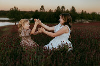 Two young girls sit in a field of red flowers at sunset, playing a hand-clapping game. One wears a white dress, the other a floral dress with a matching headband. Trees and a lake frame the background. Captured by a family photographer in Macon, GA, this moment is pure magic.