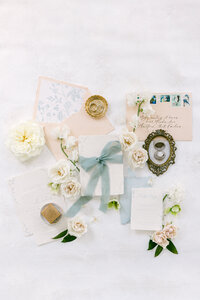 sage green and cream white wedding invitation suite flatlay with peach color rose and blush ring box