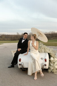 Wedding Couple Sits on Car with Umbrella