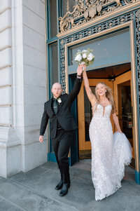 Bride and Groom happily exit the San Francisco City Hall doors while holding the brides bouquet up in the air. 
