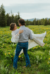 A man is carrying his soon to be bride away from the camera so that you see the back of his head. She is wearing her wedding dress. They are walking through yellow flower field.
