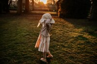  a little girl with curly hair wearing a sun hat, walking across a park at sunset with trees silhouetted in the background.