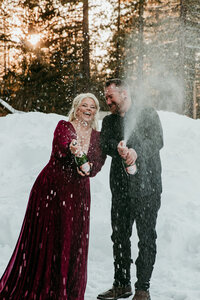 Bride in a deep red gown and groom popping champagne in a snowy forest near Lake Tahoe, celebrating their winter elopement with joy and laughter.
