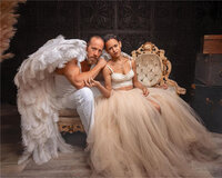 woman in a top and cream tulle sitting on a royal chair beside a man in white with white angel-like wings
