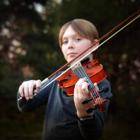 Young child playing a violin outdoors with trees.