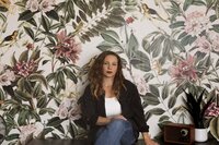 A woman with long hair sits against vibrant, floral-patterned wallpaper. She wears a black jacket, white shirt, and jeans. Next to her, a plant and vintage radio rest—she appears ready to become a white label designer in style.