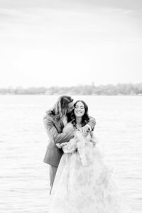 Groom kissing bride on the cheek while snuggle together beside a lake, photo is in black and white